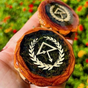 May include: Two round wooden pendants with a dark green and black resin center. Each pendant features a white laurel wreath and a stylized arrow symbol. The wood has a natural, warm brown color, and the resin has a glossy finish.