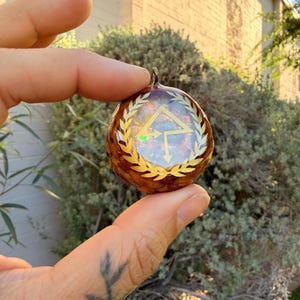 May include: A round, brown pendant with a gold-colored emblem featuring a triangle and arrows, surrounded by a laurel wreath design. The pendant has a small loop at the top for hanging. The background is blurred, with green foliage and a building.