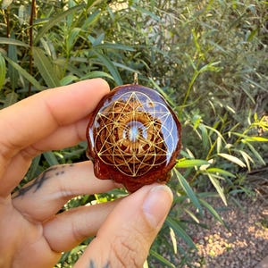 May include: A brown pendant with a gold geometric design, held against a backdrop of green foliage. The pendant has a glossy, resin-like finish and a small gold loop for hanging. The design features a detailed, symmetrical pattern.
