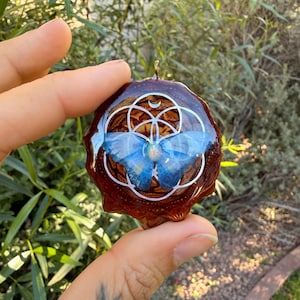 May include: A round, brown pendant with a blue butterfly design. The pendant has a silver-colored bail and a white geometric pattern. The butterfly has blue wings and a yellow center. The pendant is held up against a background of green foliage.