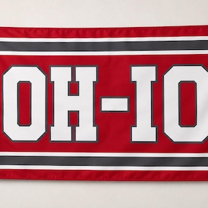 May include: A rectangular flag featuring a red background with the white text OH-IO centered in a block font. The design includes horizontal stripes in white and dark gray along the top and bottom edges, with metal grommets visible on the left side.