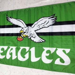 May include: Green flag featuring a white eagle clutching a football in its talons. The eagle is soaring above three black and white stripes. The words "EAGLES'" are displayed in white lettering beneath the eagle.