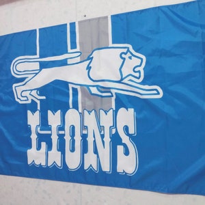 May include: A blue and white flag featuring a white lion running to the right, with the word "LIONS" in white lettering below the lion.