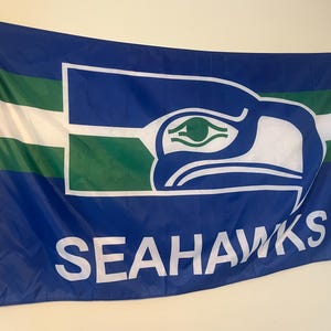 Puede incluir: Bandera azul con el logotipo de los Seattle Seahawks. La bandera presenta una cabeza de halcón estilizada en blanco y verde sobre un fondo azul, con la palabra "SEAHAWKS" en letras blancas.