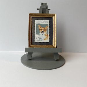 May include: A framed vintage postage stamp featuring a dog with a light brown and white coat. The stamp is from Cambodia and features the text "Royaume du Cambodge" and "1500R". The stamp is displayed on a small wooden easel.