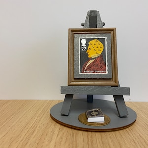 Puede incluir: Un caballete en miniatura con un sello enmarcado que presenta un retrato de Charles Babbage, considerado el padre de la computadora. El sello es del Reino Unido y presenta el texto "Babbage - Computer" y "22". Un pequeño libro se encuentra en la base del caballete.