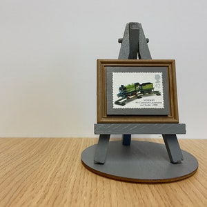 May include: A postage stamp featuring a green Hornby M1 clockwork locomotive and tender from 1948, displayed in a small brown frame mounted on a grey wooden easel. The stamp includes the text Hornby M1 Clockwork Locomotive and Tender c.1948.