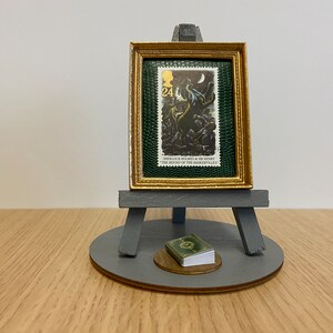 Può includere: Un cavalletto in miniatura con un francobollo incorniciato che presenta un'illustrazione di un cane da caccia dalla storia di Sherlock Holmes "Il Segugio dei Baskerville". Un piccolo libro verde si trova alla base del cavalletto.
