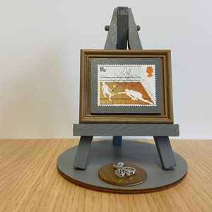 May include: A small grey wooden easel holding a framed postage stamp featuring a squash game illustration with the text 11p. A small silver-toned metal tennis racket charm is attached to the circular grey base of the easel stand.