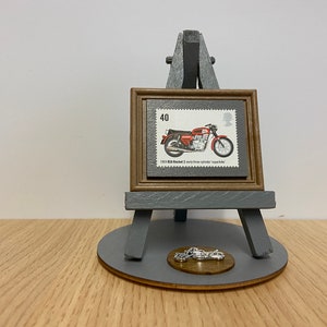 May include: A miniature easel displaying a framed vintage stamp featuring a red 1963 BSA Rocket 3 motorcycle. The stamp is on a grey background with the text "40" and "1963 BSA Rocket 3 early three cylinder superbike". A small silver motorcycle charm is on the base of the easel.