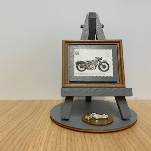 May include: A framed vintage stamp featuring a 1930 Brough Superior bespoke luxury motorcycle. The stamp is displayed on a small wooden easel with a gray base and a round wooden base.