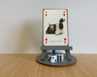 May include: A playing card featuring an illustration of a black and white American Cocker Spaniel, labelled with the number 4 and red heart symbols. The card rests on a small grey wooden easel stand, accompanied by two white six-sided dice.