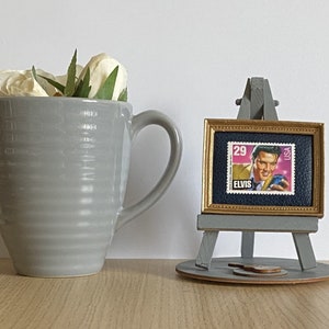 May include: A grey ceramic mug with white flowers inside, next to a miniature easel with a framed Elvis Presley postage stamp. The stamp is blue and features a portrait of Elvis Presley in a gold-trimmed frame.