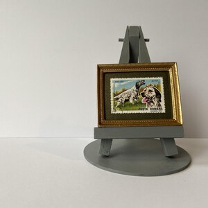 Puede incluir: Un sello postal rumano vintage enmarcado con un perro de caza con el texto "POSTA ROMANA" en un marco dorado sobre un caballete gris.