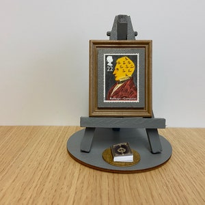 May include: A miniature wooden easel in grey holds a framed postage stamp featuring a profile of Charles Babbage with the text Babbage - Computer and the number 22. A tiny book rests on the circular grey base in front of the easel.