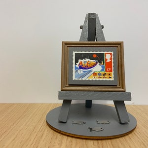 May include: A framed vintage postage stamp featuring a lifeboat with a red flare in the sky. The stamp is on a gray background with a brown frame and is displayed on a gray easel with a round base. Three silver fish shapes are on the base.