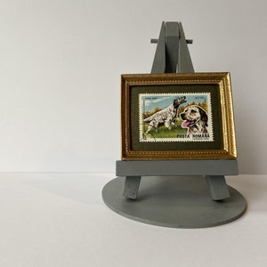 Puede incluir: Un sello postal rumano vintage enmarcado con un perro de caza con el texto "POSTA ROMANA" en un marco dorado sobre un caballete gris.