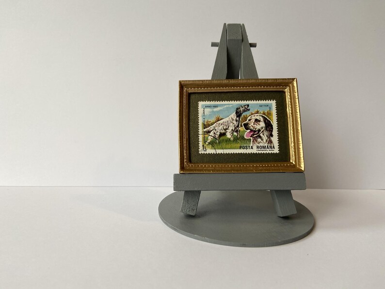 Puede incluir: Un sello postal rumano vintage enmarcado con un perro de caza con el texto "POSTA ROMANA" en un marco dorado sobre un caballete gris.