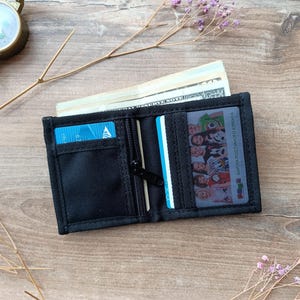 Könnte beinhalten: Geöffnete schwarze Geldbörse mit Reißverschluss, gefüllt mit Bargeld und Kreditkarten. Die Geldbörse liegt auf einer Holzoberfläche, im Hintergrund ein Kompass und Zweige.