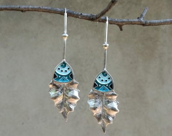 May include: A pair of silver dangle earrings with a leaf design. The leaves are detailed and textured. The top of each earring features a blue, white, and black geometric design.