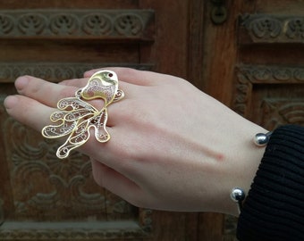 May include: A gold and silver filigree ring shaped like a fish. The ring is worn on a person's finger. The ring is on a background of a wooden door.