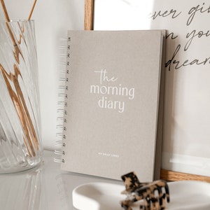 Könnte beinhalten: Ein beigefarbenes Spiralnotizbuch mit dem Text "the morning diary" und "MY DAILY LINES" auf dem Einband.