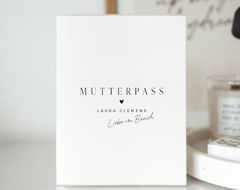 May include: White card with black text that reads "Mutterpass" with a small heart symbol below. Below the text is "Laura Clemens" and "Liebe im Bauch" in cursive.