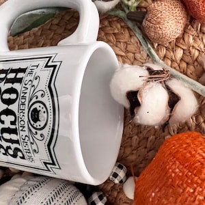 May include: White ceramic mug with a black and white graphic of a label that reads "Hocus Pocus The Sanderson Sisters". The mug is sitting on a woven surface with cotton bolls and other autumnal decor.