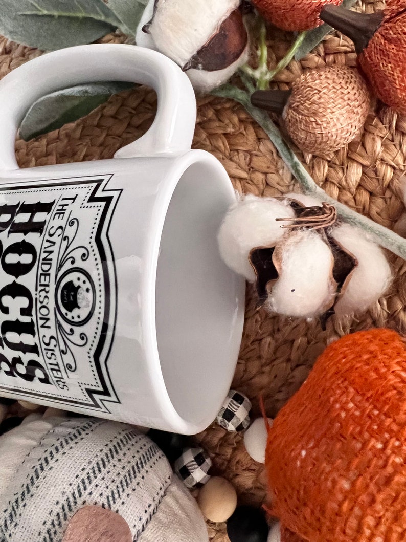 May include: White ceramic mug with a black and white graphic of a label that reads "Hocus Pocus The Sanderson Sisters". The mug is sitting on a woven surface with cotton bolls and other autumnal decor.
