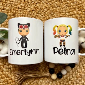 May include: Two white ceramic mugs with black handles. The mug on the left has a cartoon image of a girl dressed as Catwoman with the name "Emerlynn" printed below. The mug on the right has a cartoon image of a girl dressed as Harley Quinn with the name "Petra" printed below.