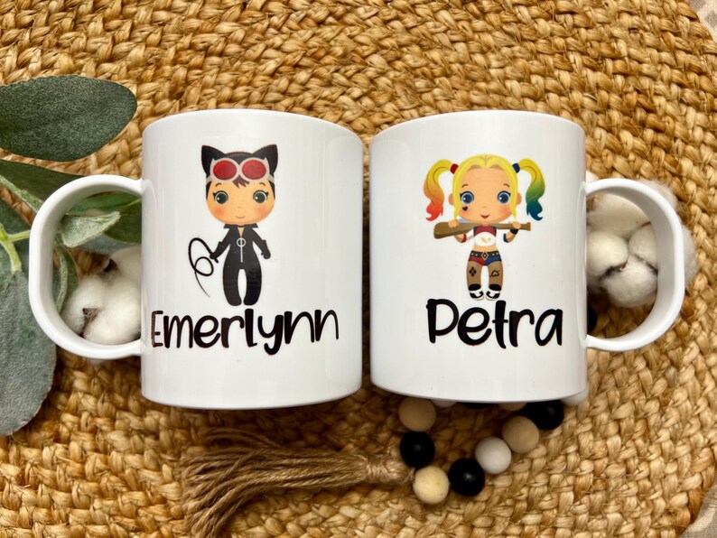 May include: Two white ceramic mugs with black handles. The mug on the left has a cartoon image of a girl dressed as Catwoman with the name "Emerlynn" printed below. The mug on the right has a cartoon image of a girl dressed as Harley Quinn with the name "Petra" printed below.