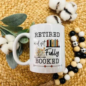 May include: White ceramic mug with a black and white design that says "Retired and yet Fully Booked". The design includes a stack of books, a book with flowers, and an arrow.
