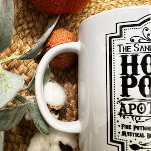 May include: White ceramic mug with a black and white graphic design that says "The Sanderson Sisters'  Hot  Potion Apothecary" with the text "Fine Potions & Mystical Brews" at the bottom.
