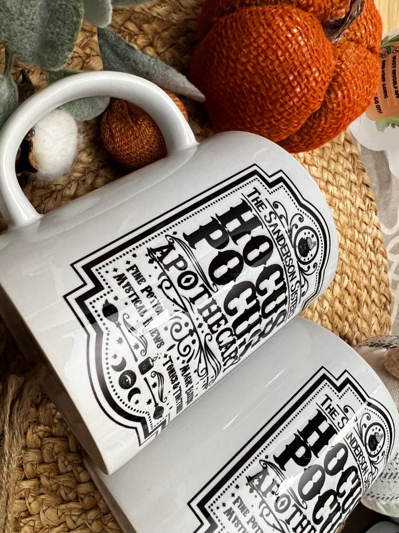 May include: White ceramic mug with a black and white graphic design featuring the text "The Sanderson Sister's Hocus Pocus Apothecary" and a witch's cauldron. The design includes stars, moons, and other witchy symbols.