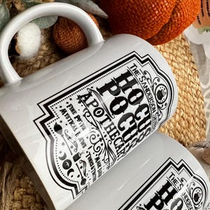 May include: White ceramic mug with a black and white graphic design featuring the text "The Sanderson Sister's Hocus Pocus Apothecary" and a witch's cauldron. The design includes stars, moons, and other witchy symbols.