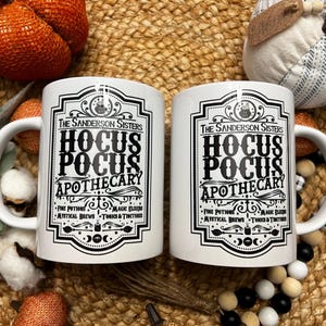 May include: Two white ceramic mugs with black text and graphics. The text reads "The Sanderson Sisters Hocus Pocus Apothecary" with a list of ingredients below.