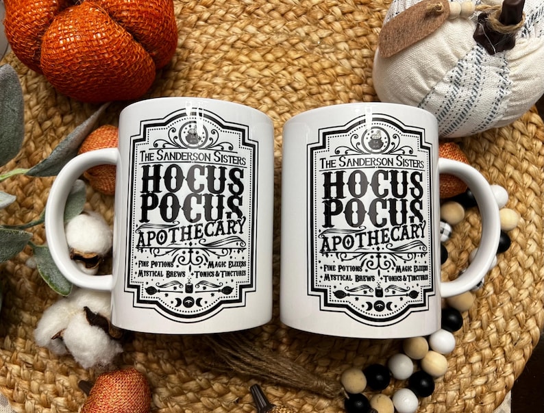 May include: Two white ceramic mugs with black text and graphics. The text reads "The Sanderson Sisters Hocus Pocus Apothecary" with a list of ingredients below.
