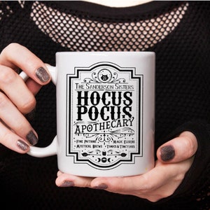 May include: White ceramic mug with a black and white graphic design featuring the text "The Sanderson Sisters Hocus Pocus Apothecary" with a black cat illustration. The text below reads "Fine Potions, Magic Elixirs, Mystical Brews, Tonics & Tinctures".
