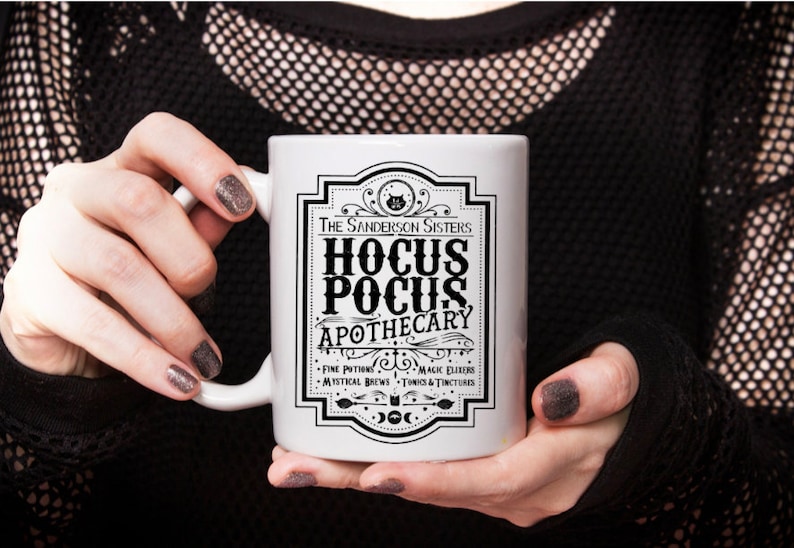 May include: White ceramic mug with a black and white graphic design featuring the text "The Sanderson Sisters Hocus Pocus Apothecary" with a black cat illustration. The text below reads "Fine Potions, Magic Elixirs, Mystical Brews, Tonics & Tinctures".