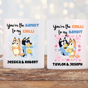May include: Two white ceramic mugs with cartoon characters and the phrases "You're the Bandit to my Chilli" and "You're the Chilli to my Bandit". The mugs have names below the text. One mug has blue accents, the other pink.
