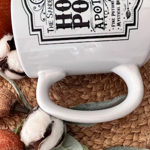 May include: White ceramic mug with a black and white graphic design that reads "The Sanders Hot Pot Apothecary Fine Potions Mystical Brews". The mug is sitting on a woven brown surface.