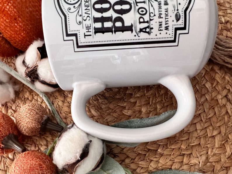 May include: White ceramic mug with a black and white graphic design that reads "The Sanders Hot Pot Apothecary Fine Potions Mystical Brews". The mug is sitting on a woven brown surface.