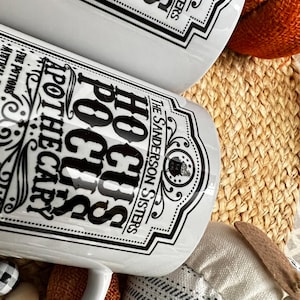 May include: Two white ceramic mugs with black lettering. The mugs feature a vintage apothecary label design with the text "Hocus Pocus Apothecary" and "The Sanderson Sisters".