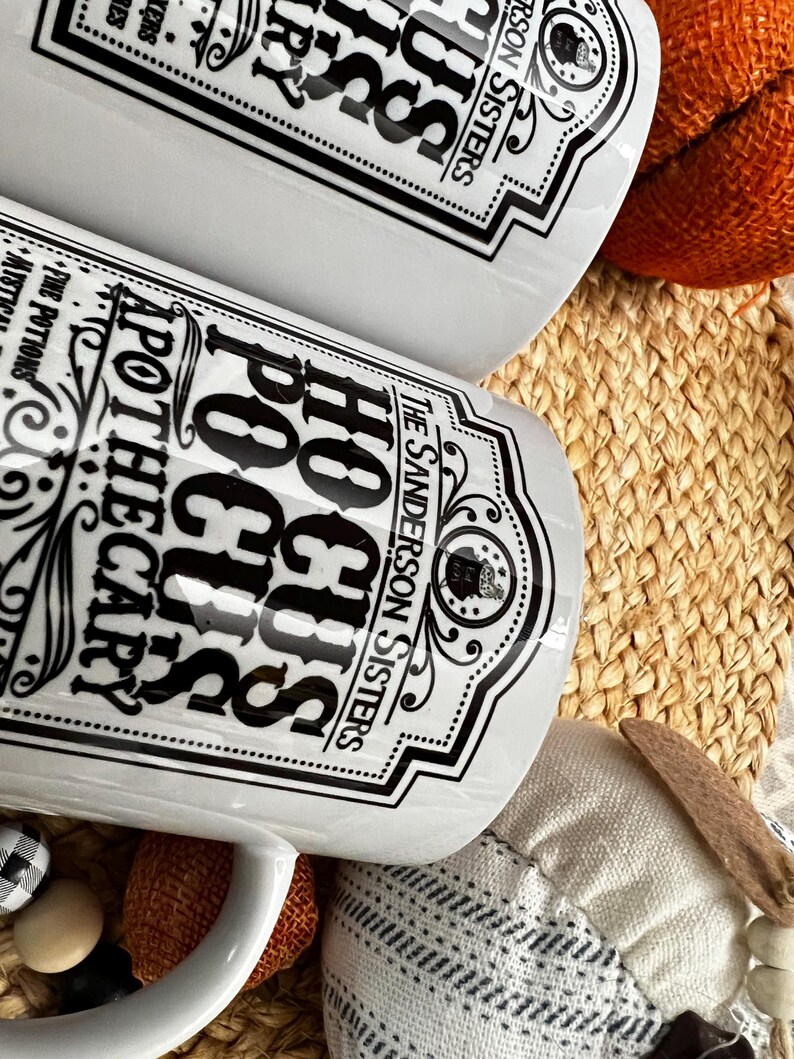 May include: Two white ceramic mugs with black lettering. The mugs feature a vintage apothecary label design with the text "Hocus Pocus Apothecary" and "The Sanderson Sisters".