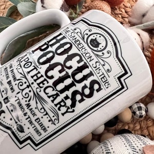 May include: White ceramic mug featuring a black and white graphic of a potion bottle label that reads "The Sanderson Sisters Hocus Pocus Apothecary" with a black cauldron and witch silhouette. The label also includes the text "The Potions, Magic Elixers, Mystical Brews, Tonics & Tinctures".