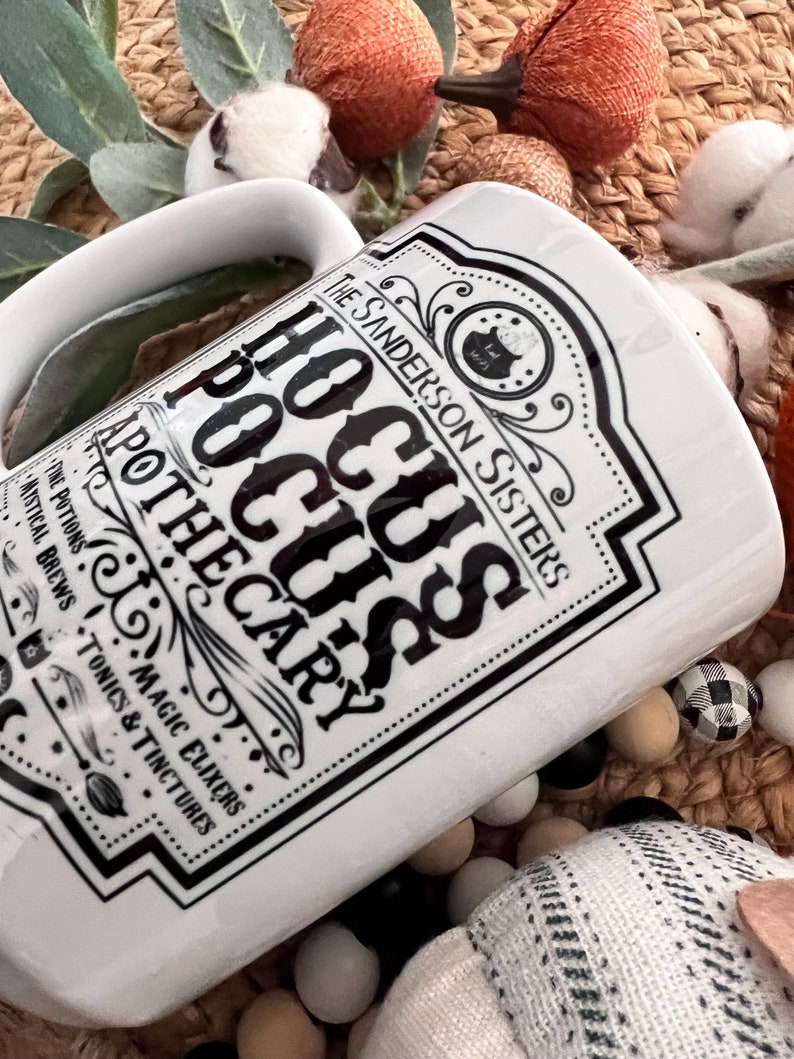 May include: White ceramic mug featuring a black and white graphic of a potion bottle label that reads "The Sanderson Sisters Hocus Pocus Apothecary" with a black cauldron and witch silhouette. The label also includes the text "The Potions, Magic Elixers, Mystical Brews, Tonics & Tinctures".