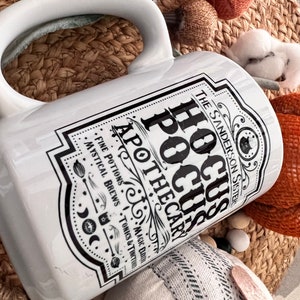 May include: White ceramic mug with a black and white graphic design featuring the text "Hocus Pocus Apothecary" and the words "Fine Potions, Mystical Brews, Tonics & Tinctures, Magic Does Exist".