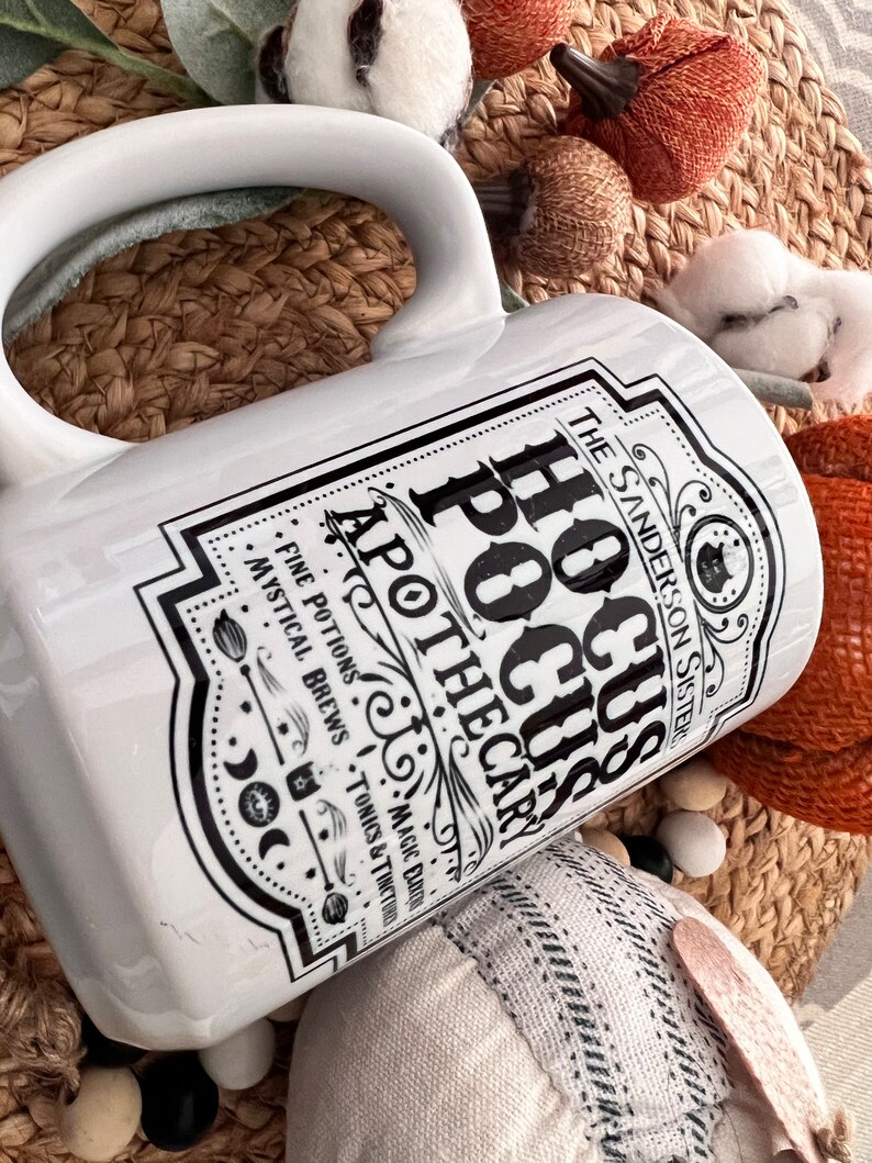 May include: White ceramic mug with a black and white graphic design featuring the text "Hocus Pocus Apothecary" and the words "Fine Potions, Mystical Brews, Tonics & Tinctures, Magic Does Exist".