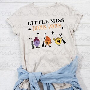 May include: A light beige t-shirt with the text "Little Miss Hocus Pocus" in black. Three cartoon characters are holding broomsticks. The characters are purple, orange, and yellow.