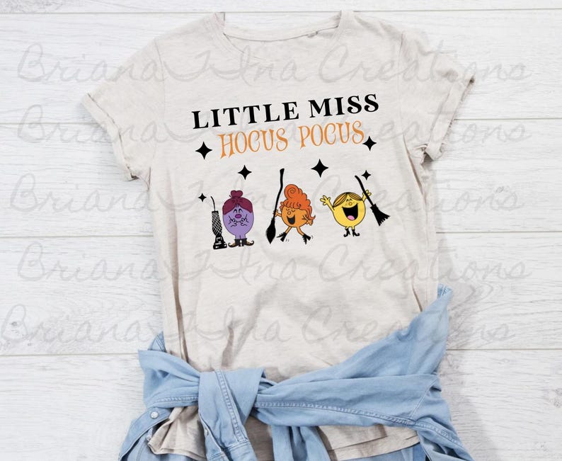 May include: A light beige t-shirt with the text "Little Miss Hocus Pocus" in black. Three cartoon characters are holding broomsticks. The characters are purple, orange, and yellow.